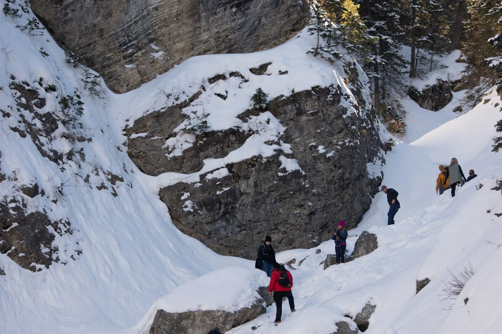 Hikers in winter clothing climbing a snowy slope below a rock face